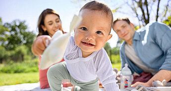 A baby reaching out with a toy, with a man and woman in the background.