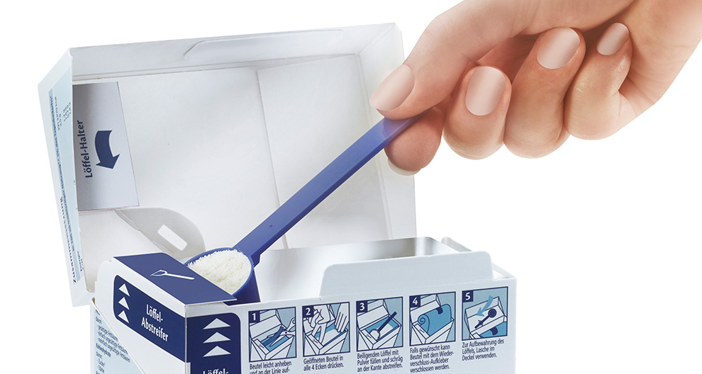 A hand is scooping a white granular substance from a blue spoon into a white box with illustrated instructions.