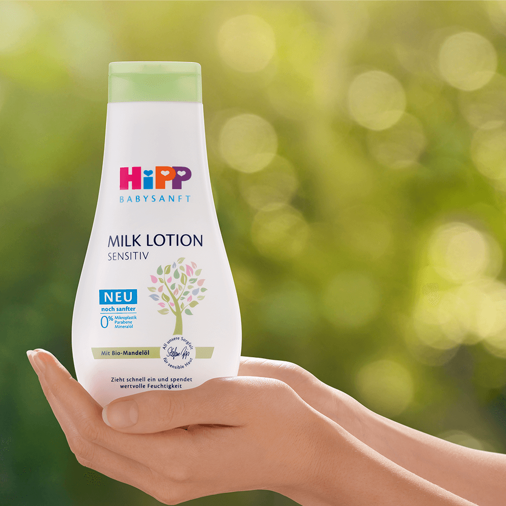 A hand holds a white bottle labeled "Hipp BabySanft Milk Lotion SENSITIV" against a green background.